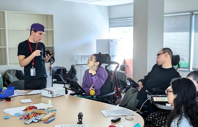 Group of people sit and stand around a table. They have AAC devices and snacks on the table