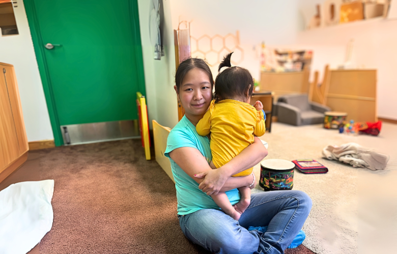 Mei-Feng sits cross legged on the floor of daycare while holding a baby