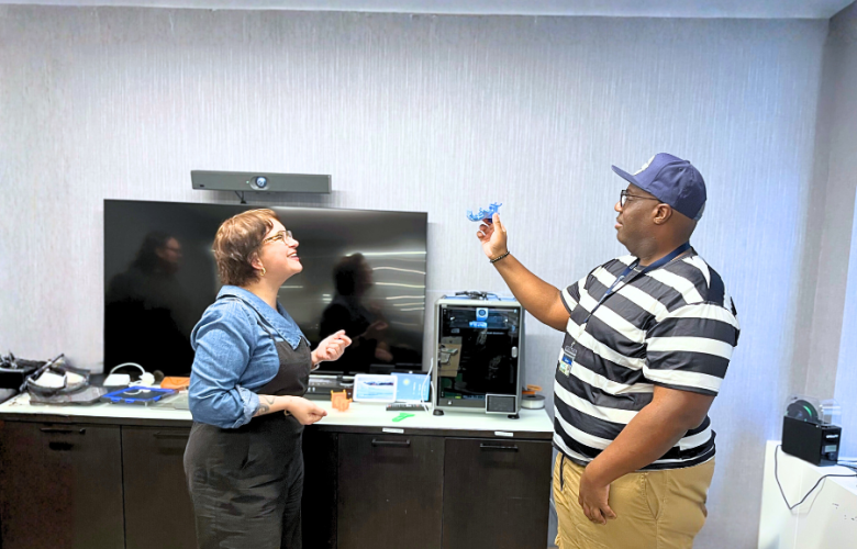 Two people stand in a room with various technological items. They are facing one another while the person on the right holds up a 3D printed item