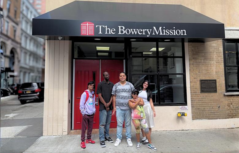 Group of people standing in front of a storefront entrance labeled “The Bowery Mission” with glass doors and brick walls on a New York City street.