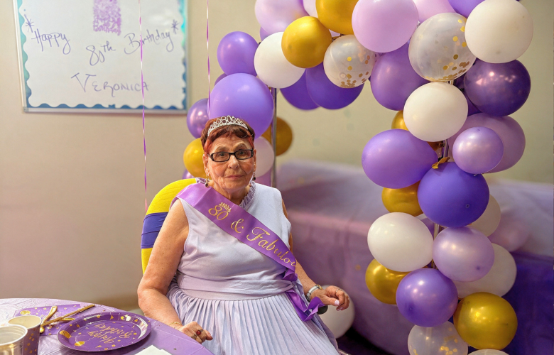 Veronica is seated indoors at her 80th birthday celebration. She is wearing a purple sash and glasses, with a large purple, white, and gold balloon cluster behind her and a hand-drawn “Happy Birthday” message on the wall.
