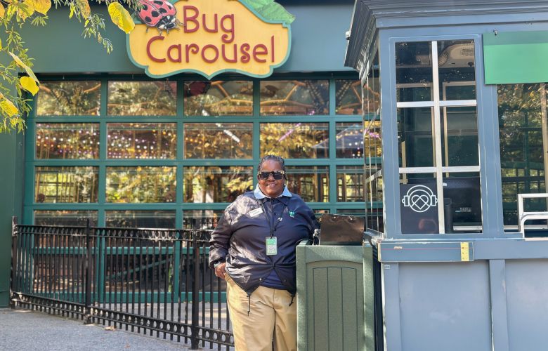 Person standing near a gray ticket booth in front of a colorful “Bug Carousel” sign, with a large glass building behind and autumn leaves framing the scene.