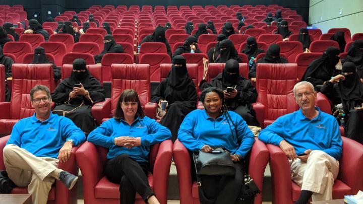 4 YAI staff sit in front of an auditorium, there are people scattered around in seats behind them, they are wearing black burkas