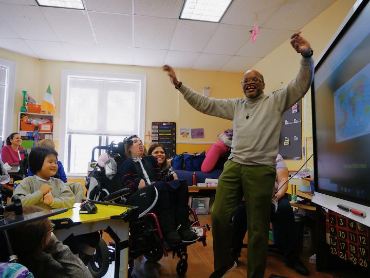 Man stands with arms in air in front of a smartboard and children and staff in classroom