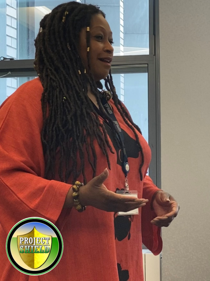 Consuelo Senior standing talking to people off camera. The project Shield logo is overlaid on the image