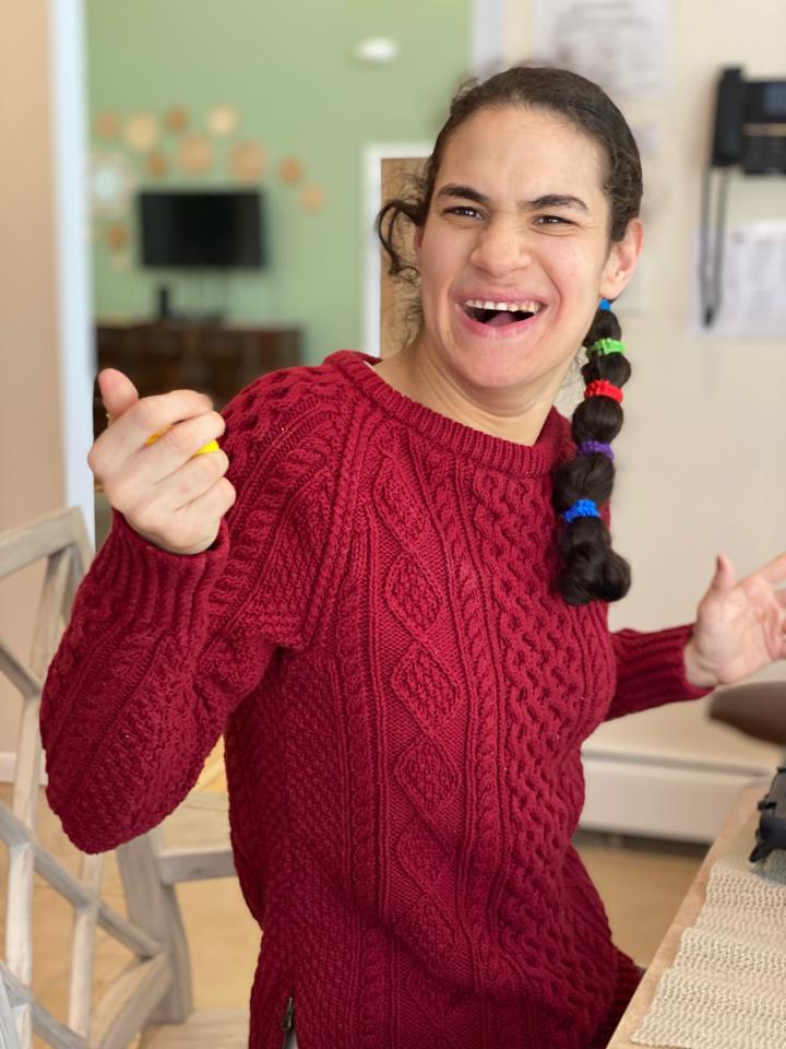 Heather wears a red sweater, has her right hand up, and is smiling