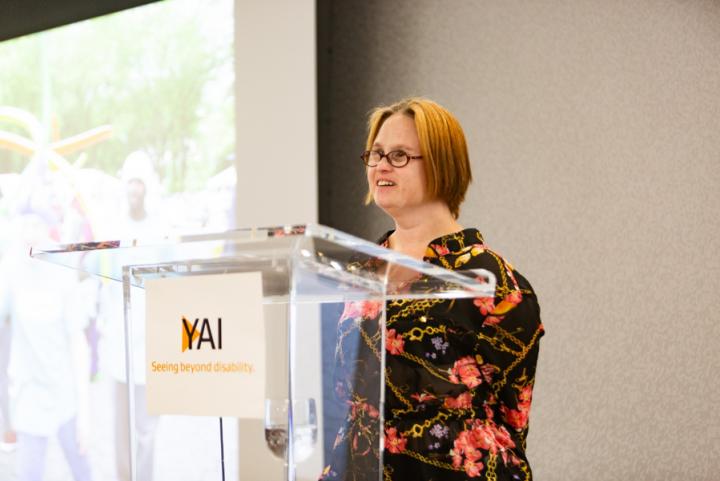 Woman in a floral dress stands at a podium, there is a sign on the front of the podium with the YAI logo on it