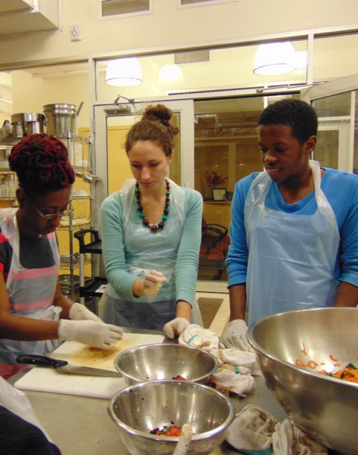 Three people stand in a kitchen, they appear to be doing food prep. There are some metal bowls in the foreground