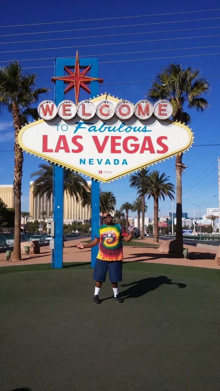 Omari wears a tie-dye shirt and stands in front of the welcome to Las Vegas sign