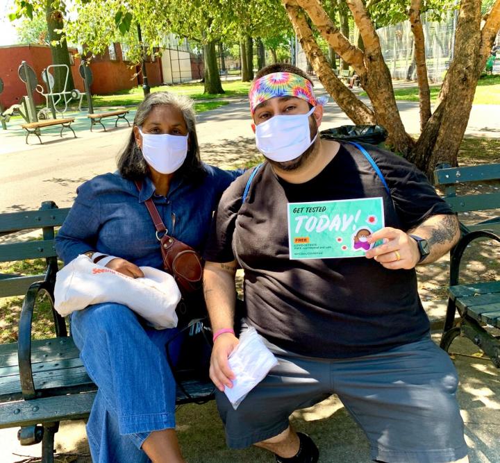 Two people wearing masks sit on a bench outside. One is holding a flyer about COVID testing