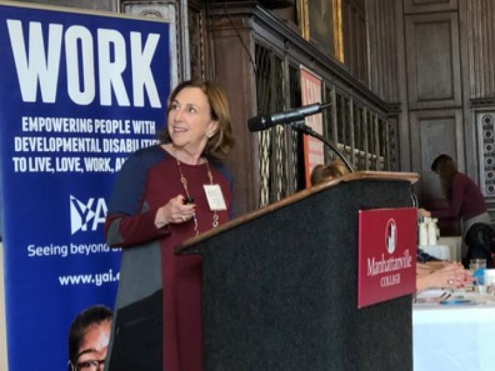 Woman standing at podium looking a little behind her. Banner behind her has the word "work" with YAI logo