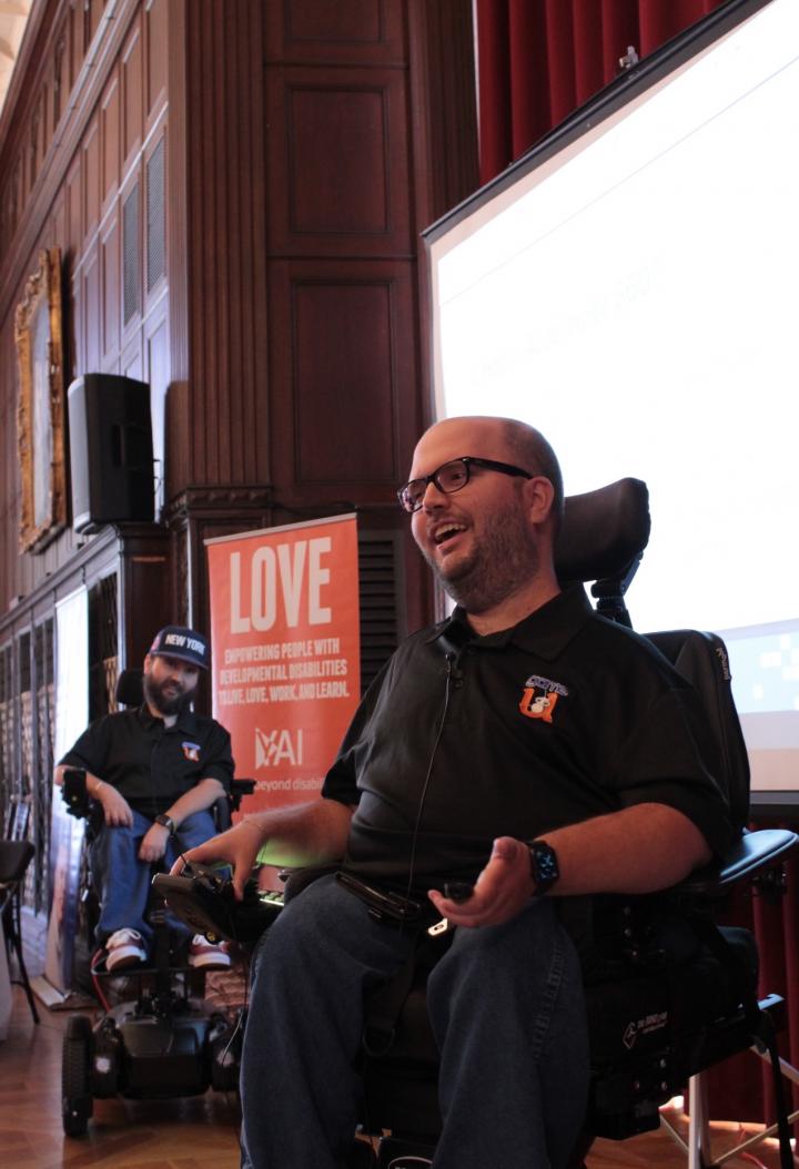 Man in wheelchair on stage, another man behind in wheelchair, both smiling