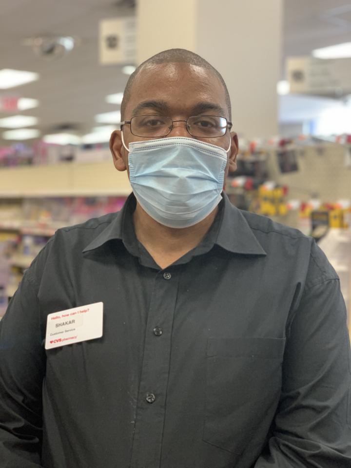 Portrait of Shakar Hollins at CVS in a shirt with a name-tag, he is wearing a disposable face mask.