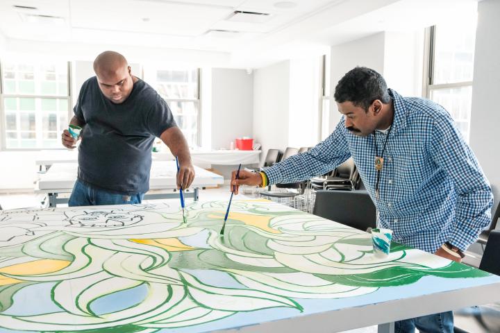 Two people painting on a large canvas placed on a table