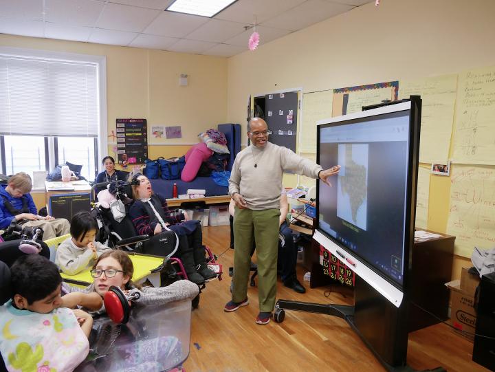 Man stands and points to a smartboard with children and staff in classroom