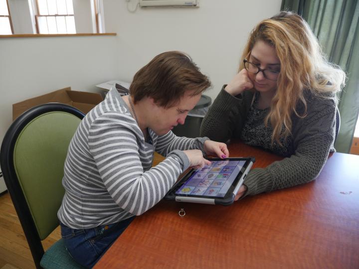 two women sit at a table, one is using an ipad while the other watches/supports