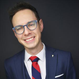 Alek Hoyos - YAI Chief of Staff Alek Hoyos wears glasses with a deep blue suit and red and blue large striped tie, he is smiling
