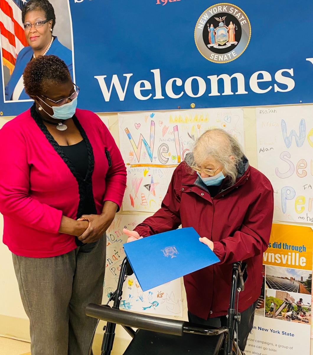 Two women wearing masks standing in front of a sign that reads "Welcomes.." woman on right is using a walking and holding a blue folder. Woman on left looks down at her.