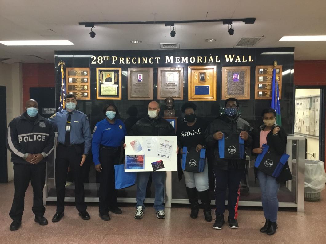 A group of people with police officers pose in a lobby with 28th precinct written on the wall