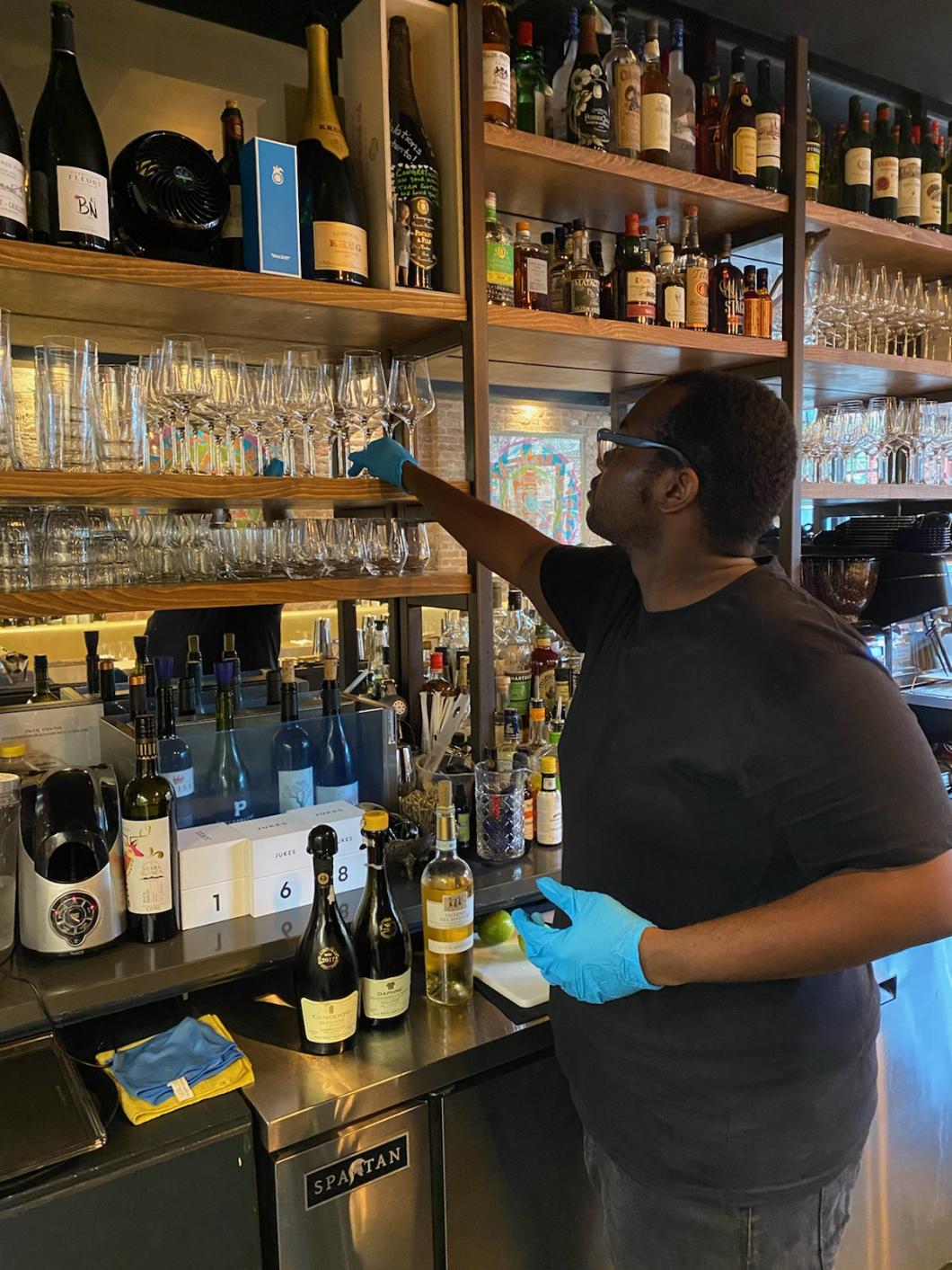 Jawann is placing a clean wine glass on a shelf. There are several bottles and glasses on the shelf.