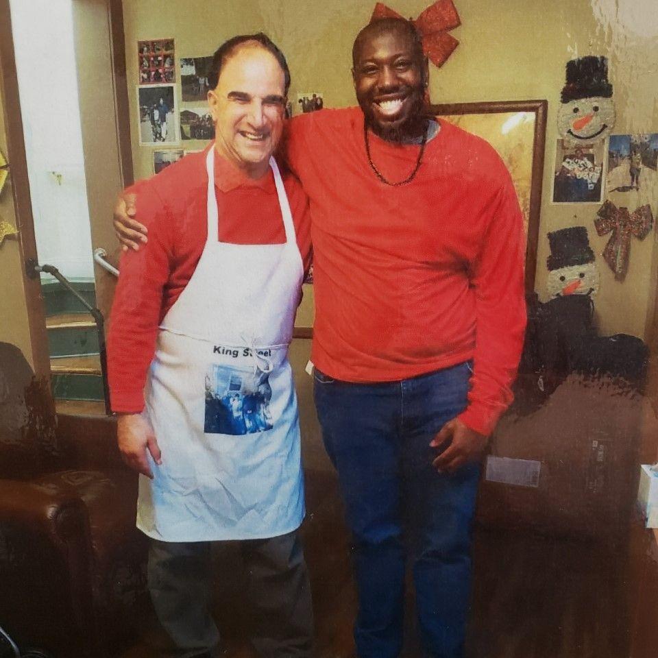Scan of a photo of Matthew Roselli (L) and Leon Owens (R) both wearing red tops, Leon has his arm around Matthew and Matthew also has on an apron. They are both cheesing for the camera