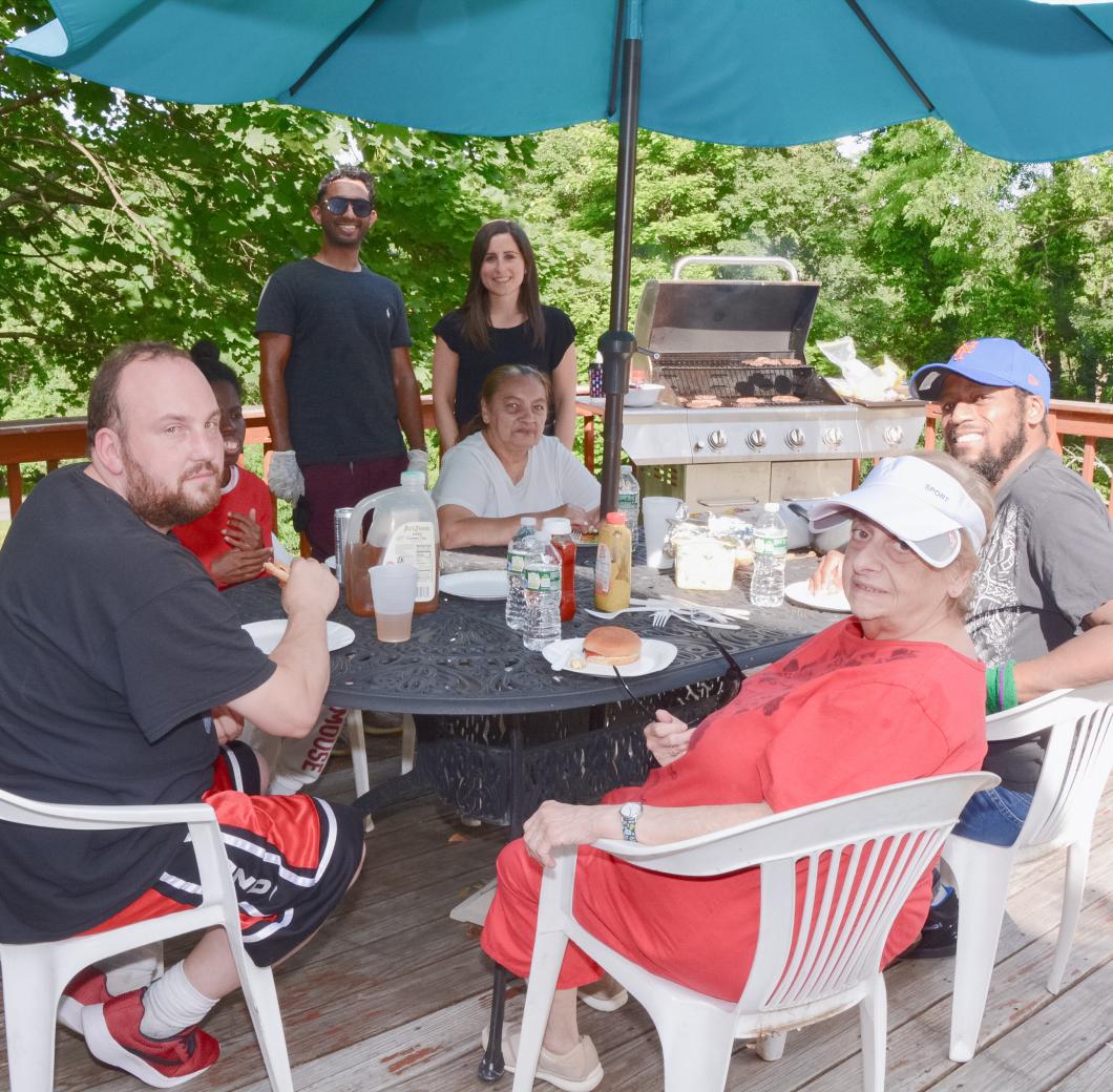 Group of people sit around a table outside underneath an umbrella, they are all turning their heads for the photograph. Two people stand in background looking on.