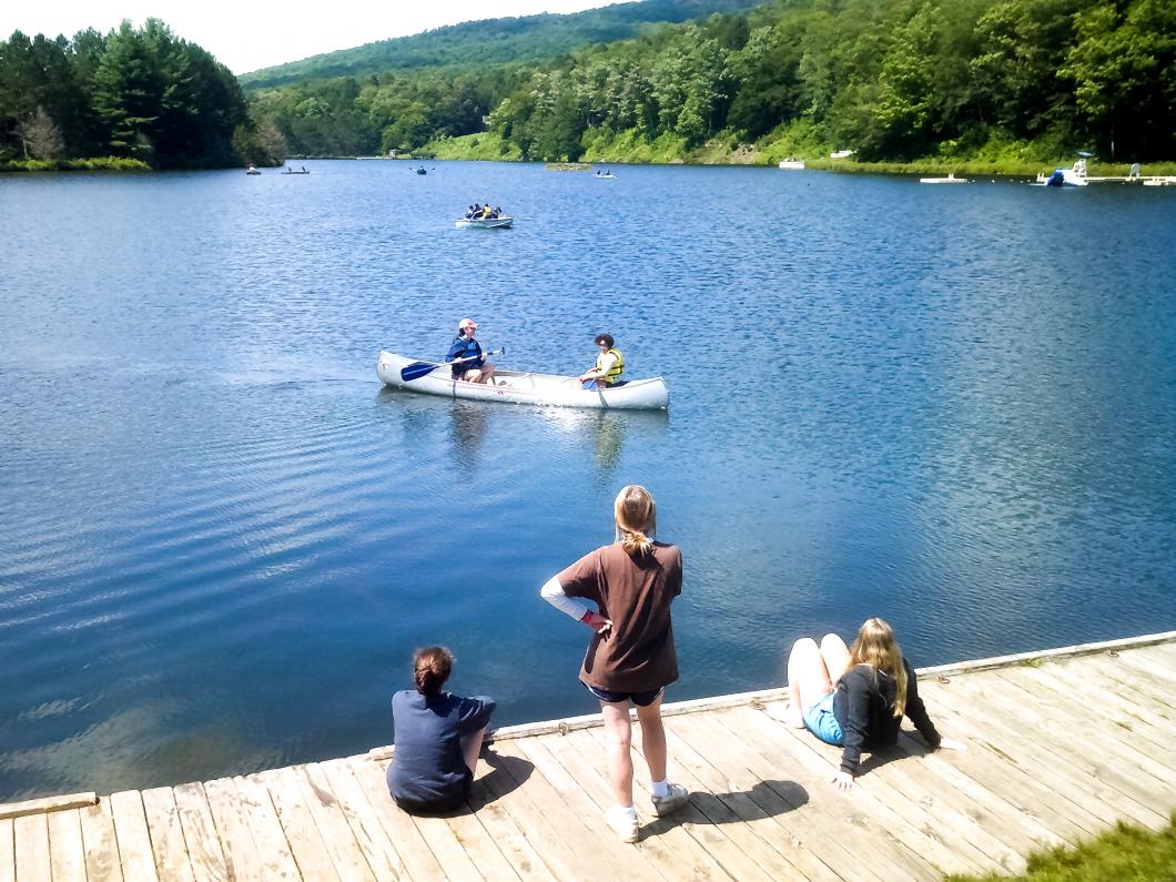 Overlooking a lake surrounded by trees, 3 people sit and stand at the jetty in the foreground. There are also people in a kayak and another boat on the lake