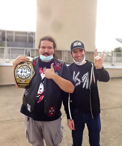 Richard and Luis stand outside the event venue, one is holding up a wrestling belt with thumb up, the other gives "devil" hand