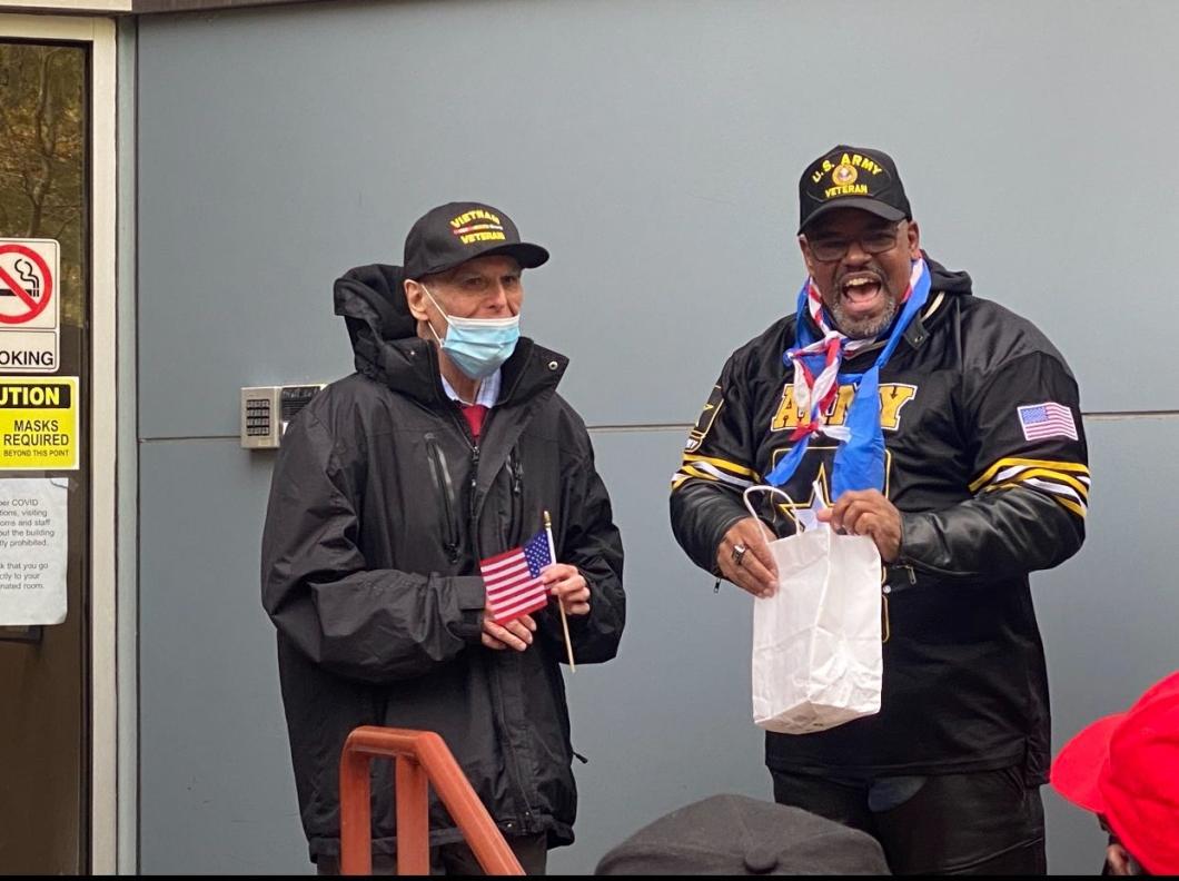 Jose and Robert stand together outside, one of them has a gift bag and is opening it and looks excited.