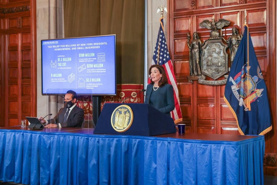 New York Governor Kathy Hochul at a press conference. Standing on a stage behind a pedastal with US and NYS flags behind her