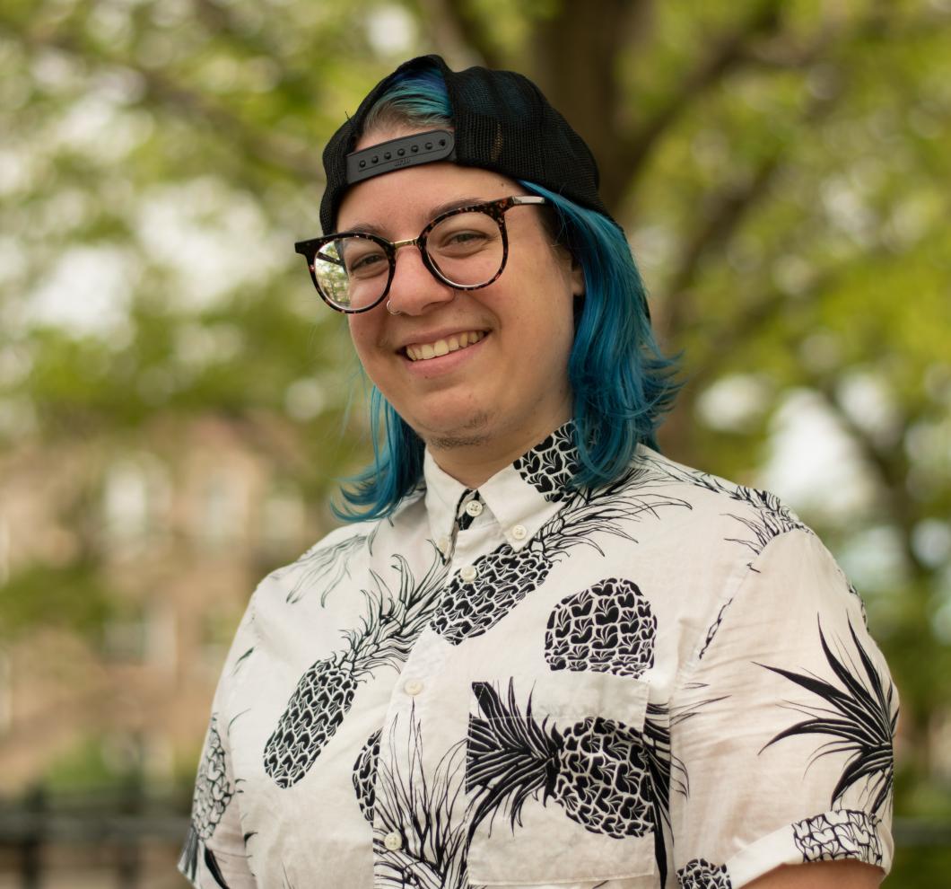 Jo Fuschetto wears glasses, backwards baseball cap, and pineapple shirt. They have teal-colored shoulder length hair.