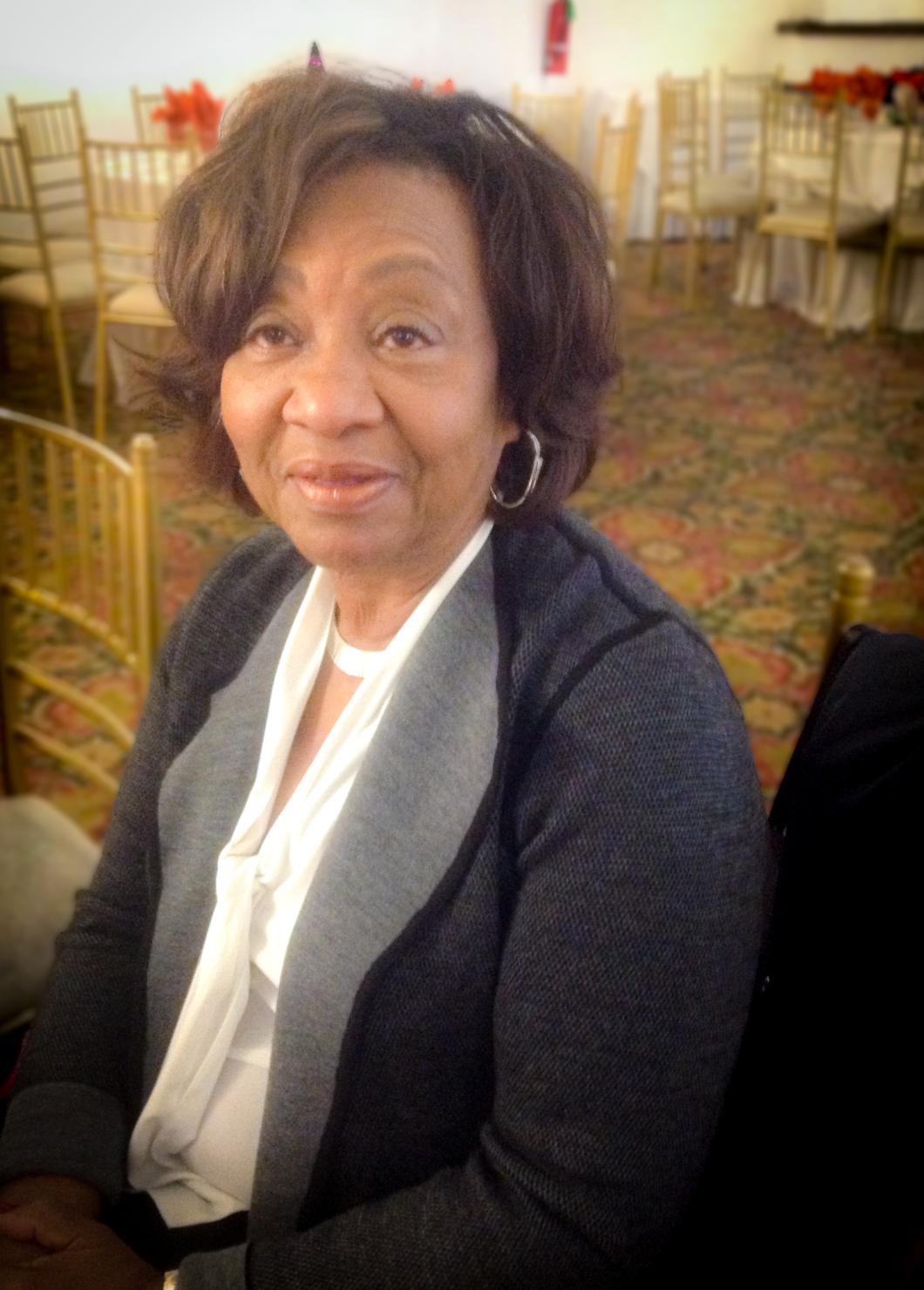 Alice Hairston sits at a chair. She wears a white shirt and blue open front top, and hoop earrings