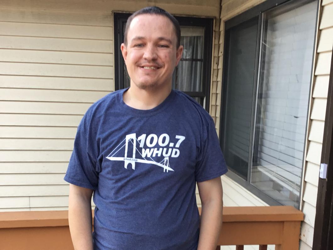Larry stands outside his house with hands in pockets. He is smiling directly at the camera and wears a 100.7 WHUD t-shirt