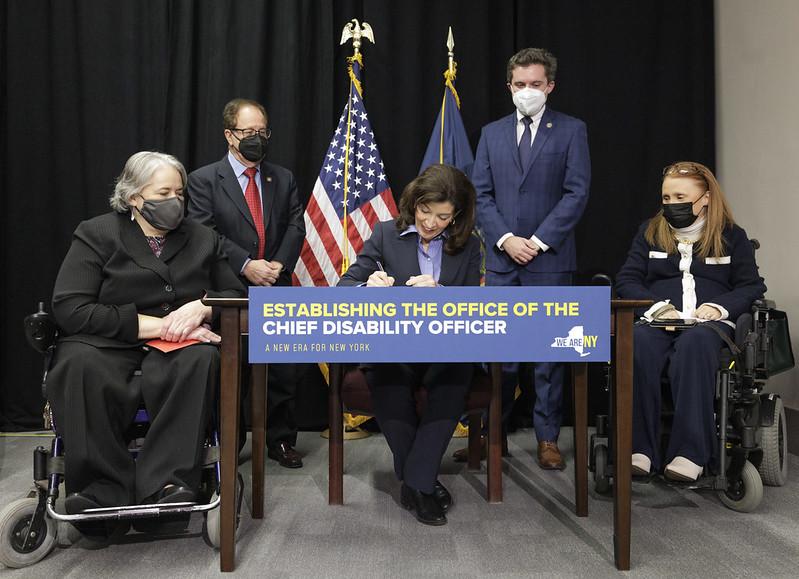 Hochul sits at a table signing a document, she has 2 people stood on either side, then further out on either side are 2 other people in wheelchairs