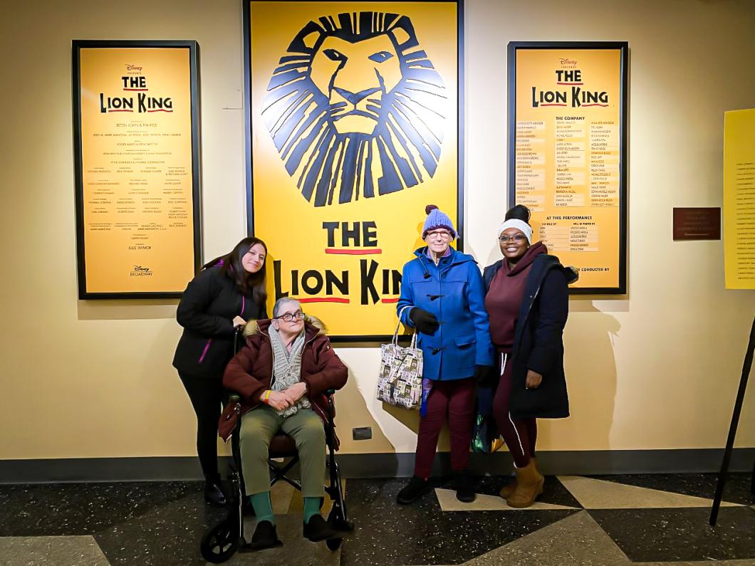 4 people pose in front of The Lion King poster. (From Left to Right) Karen Sacta, Eileen Eldred, Carole Laub, and Karla Nogais