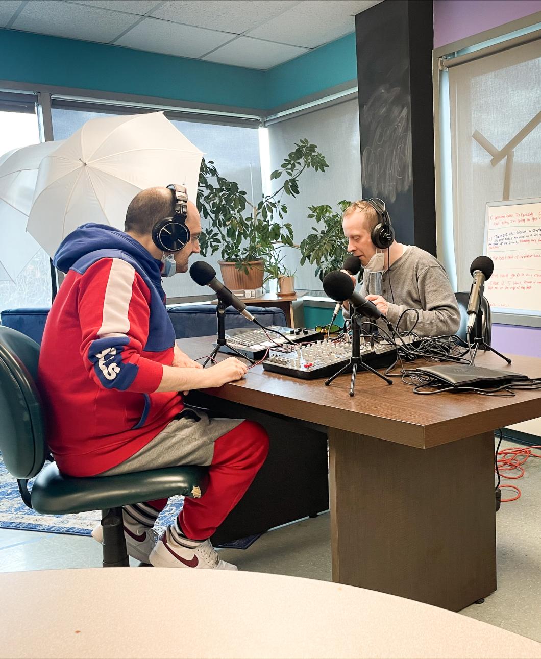 Two people sit at a desk facing one another, they are wearing headphones and using mics. There is a mixing desk between them.