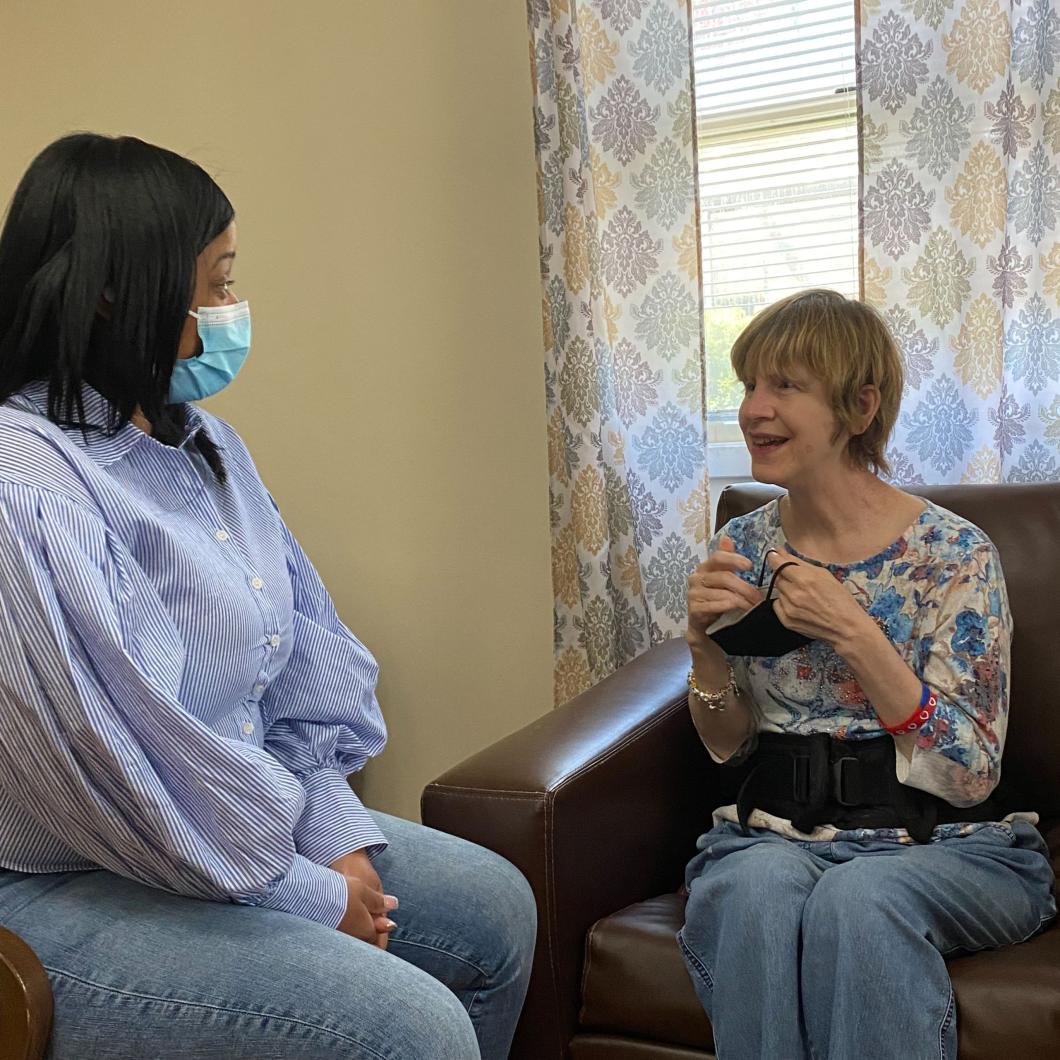 Two people sit in a home they are both in armchairs/sofa. The person on the left wears a facemask