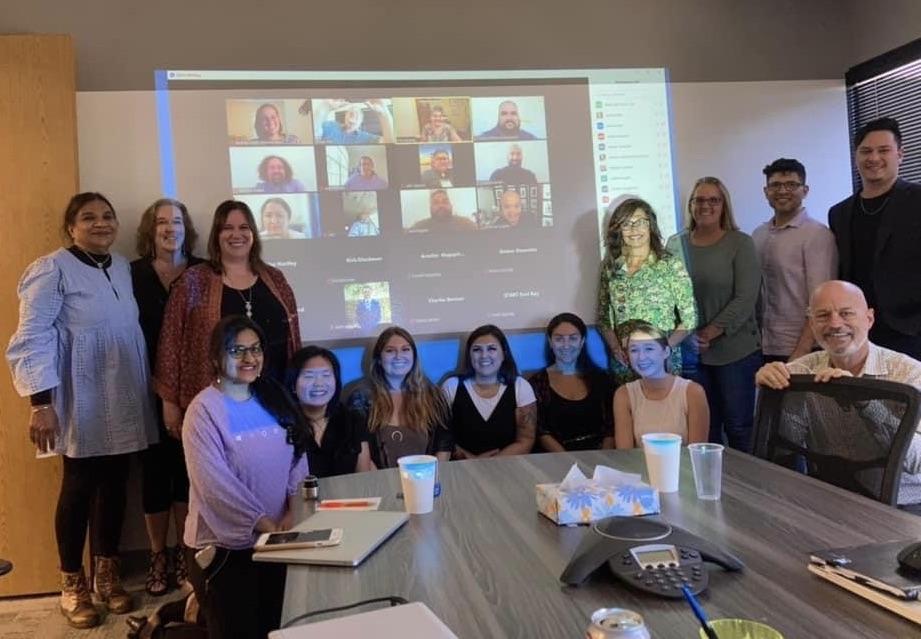 Group of 14 people gather around a screen projecting a zoom call.