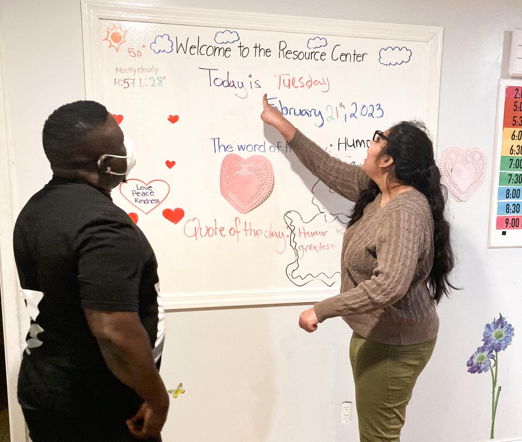 Two people stand in front of a whiteboard. Karisma is on the right and is pointing to some writing on the board