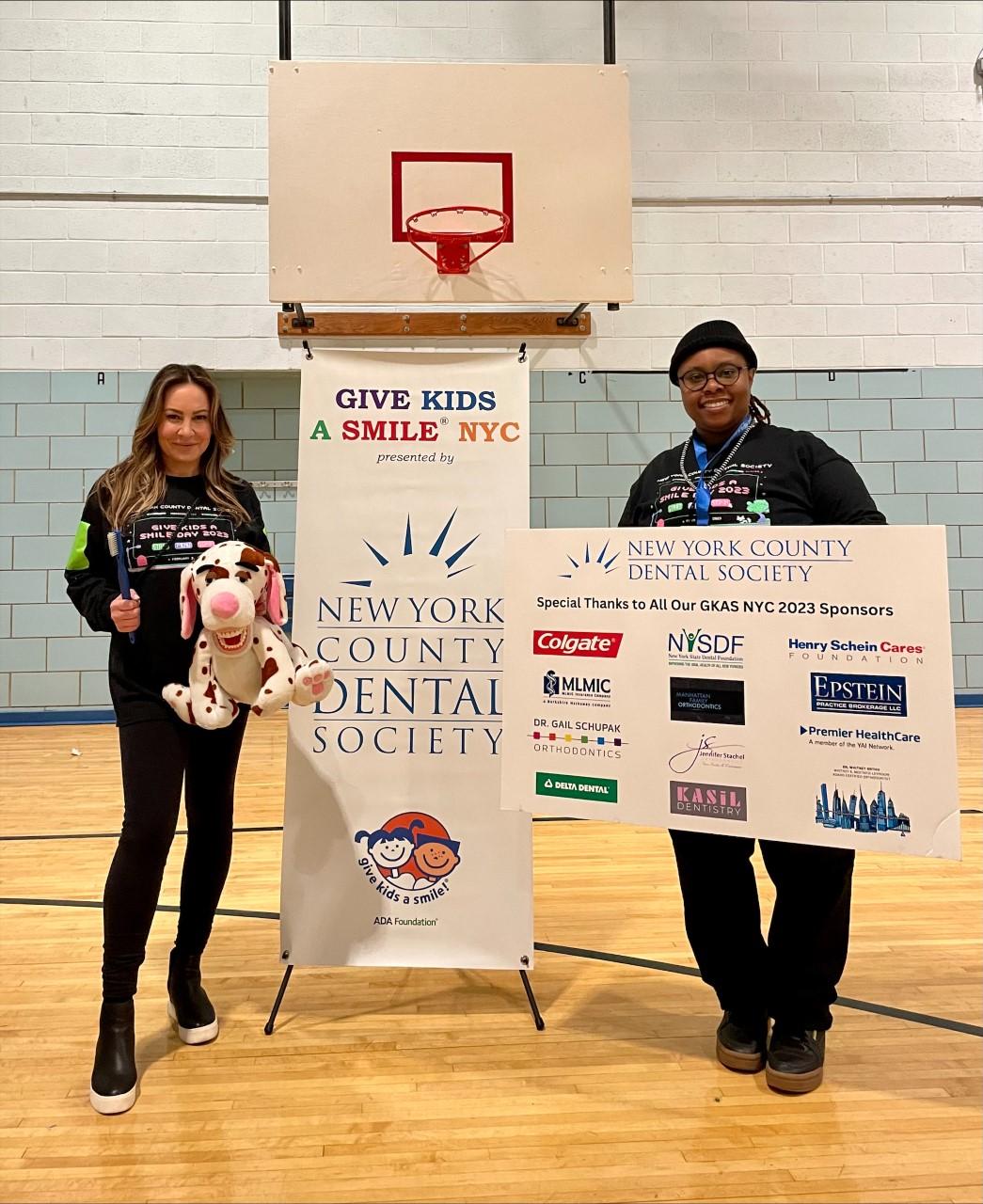 Two people stand by "Give Kids a Smile NYC" signage in a school gym