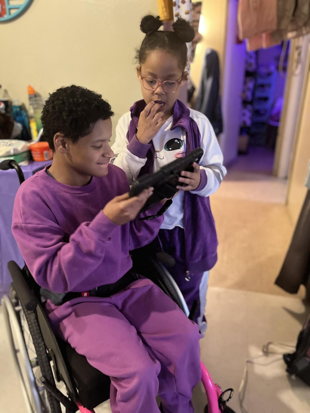 Mia (left) is in a wheelchair and holds an iPad, her sister (right) stands behind and to the side of her