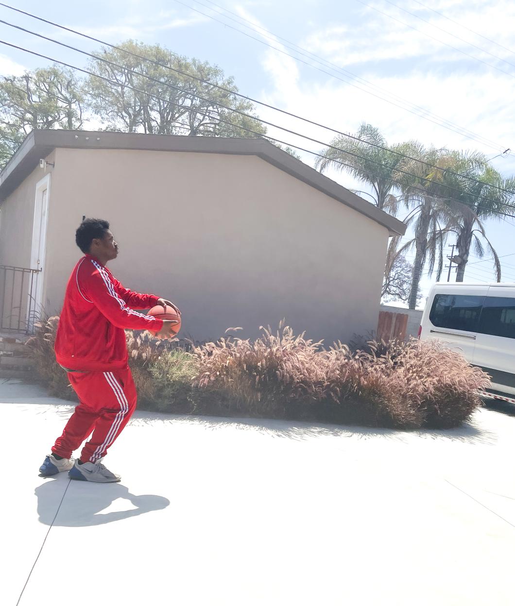 Black man in red tracksuit appears to be in motion as he holds a basketball on a driveway