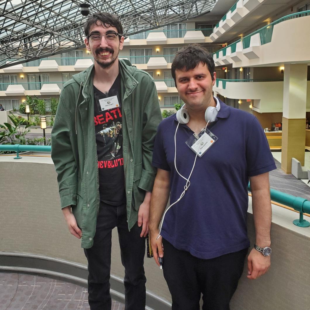 Nick and Matt stand side-by-side and pose for picture on a balcony with atrium behind them