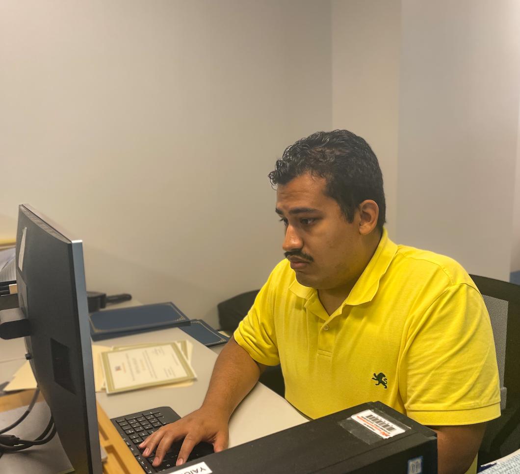 Orlando sits at a desk with a computer, he is wearing a yellow shirt and looking at the computer screen
