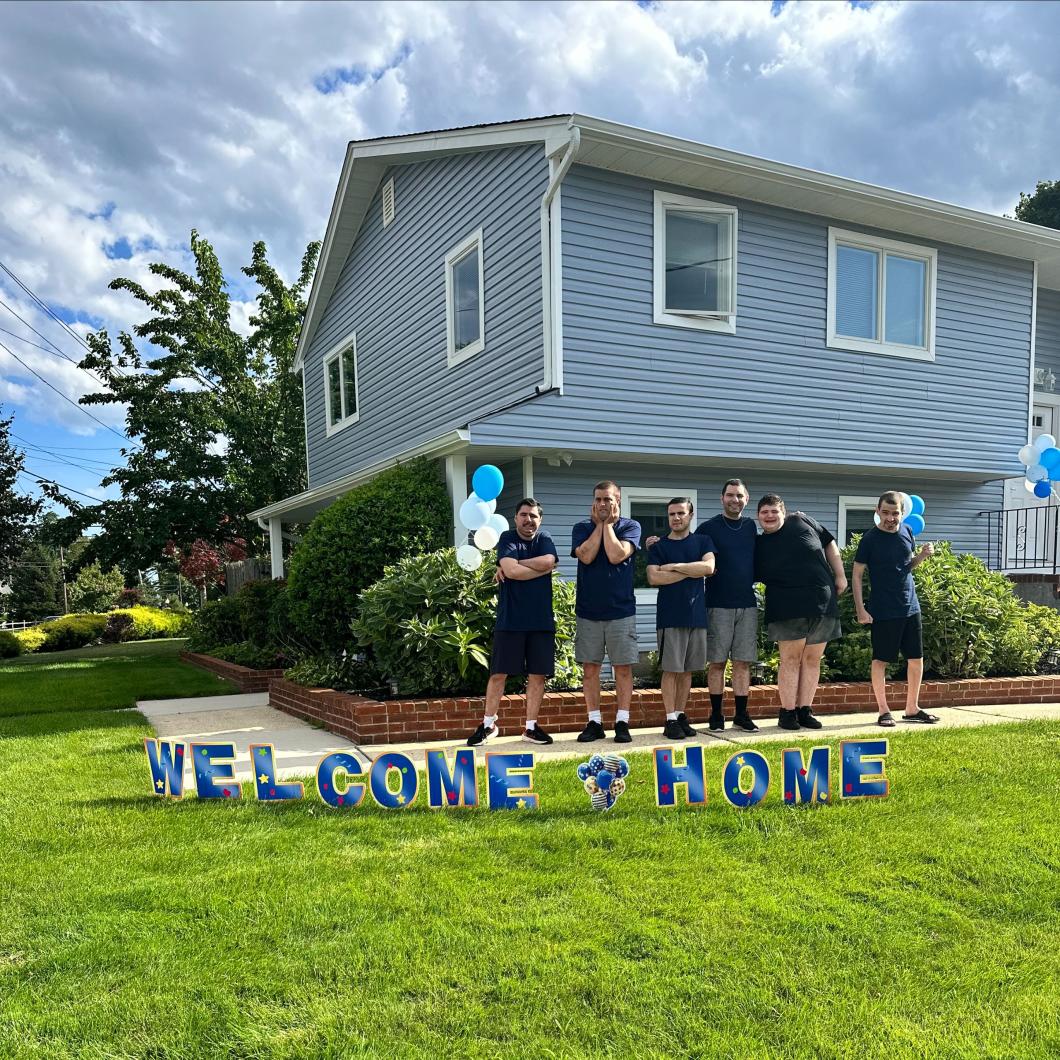 6 men stand in front of a blue house with a "welcome home" sign on the grass in front of them.