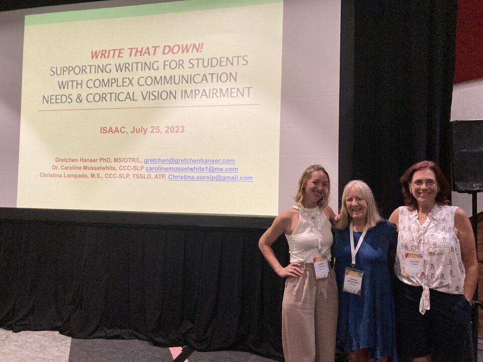 3 women stand on a stage in front of a projected image of presentation