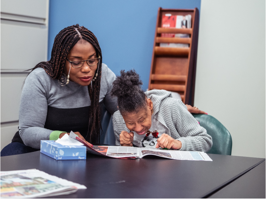 Two women sit at a table, one is supporting the other as they lean close to read something on some paper