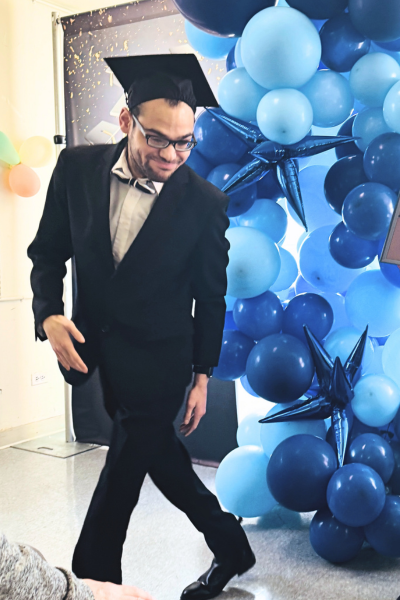 Man in suit and mortar dances by a fun collection of balloons of various shapes in various shades of blue