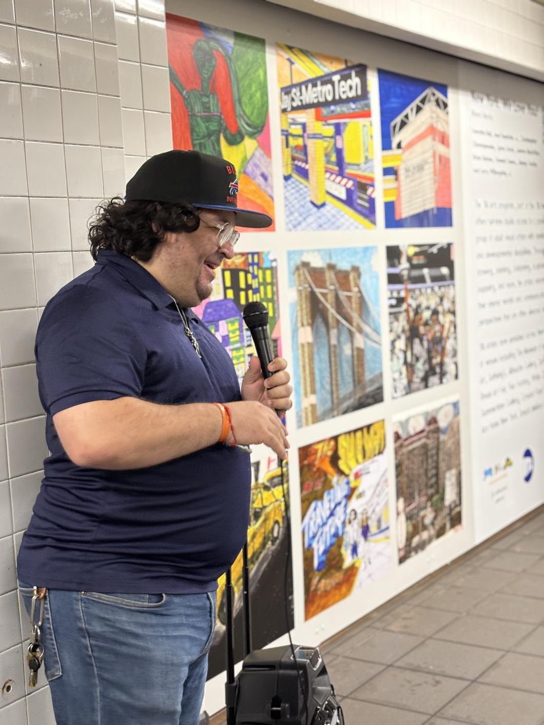 Chris stands with a mic in front of a collage of art pieces on a wall in the NY Subway
