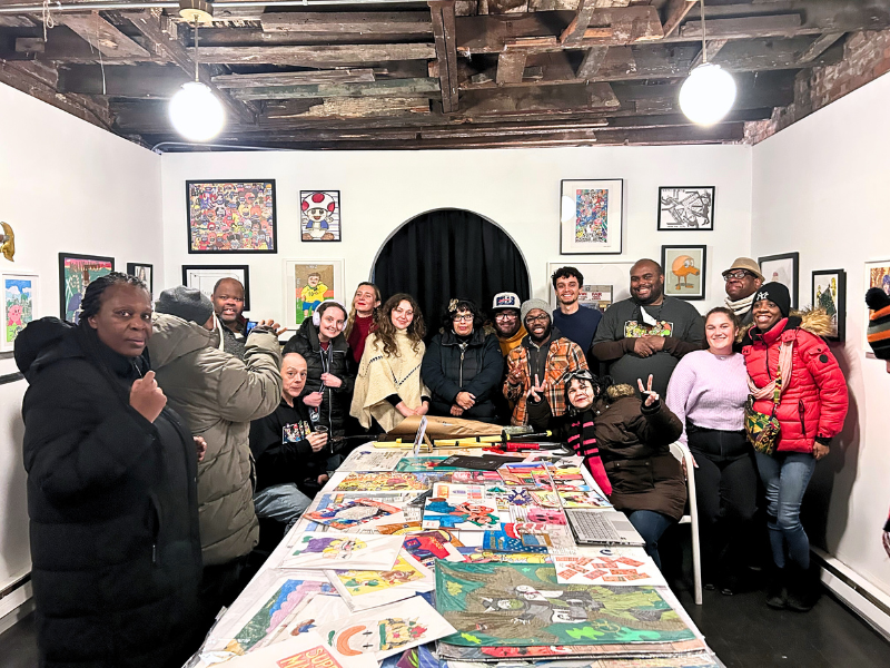 A large group of YAI Arts artists pose in a gallery. There is art on the wall as well as lots of art on a large table in front of them.